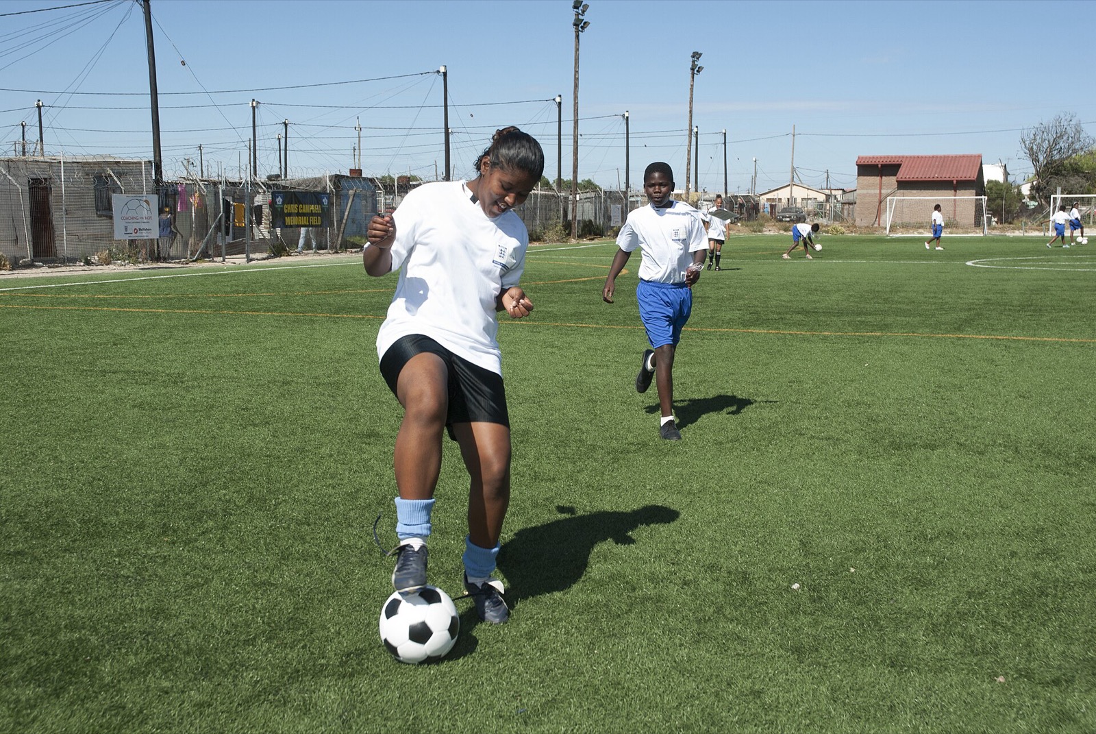 A girl dribbling a ball on a football pitch surrounded by houses in Khayelitsha